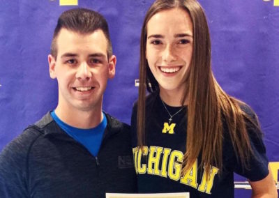 A man and woman standing next to each other, grinning with pride, holding a college signing day certificate.