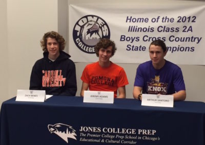 Three boys participate in a college signing day ceremony at a table in front of a banner.