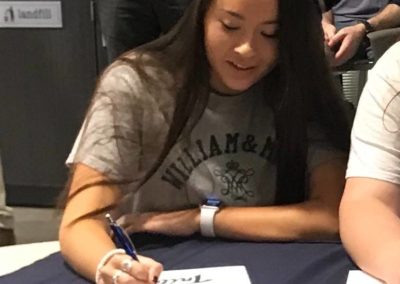 Two women participating in a college signing day event, signing papers at a table in front of a crowd.