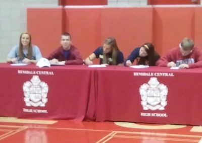 A group of people participating in a college signing day, sitting at a table and signing papers.