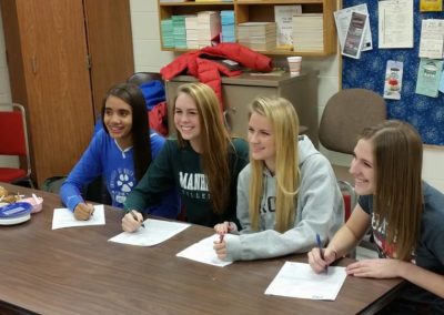 A group of girls participating in college signing day by sitting at a table and signing papers.