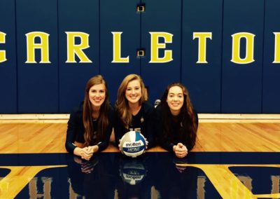 Three girls participating in a college signing day event, posing with a volleyball in front of a sign that says Carleton.
