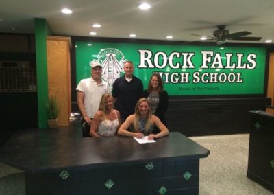 A group of people posing in front of a sign that says Rock Falls High School on college signing day.