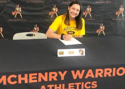 On college signing day, a girl sits at a table, eagerly signing her letter of intent.