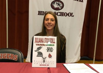 A girl participating in college signing day, sitting in front of a table with a sign.