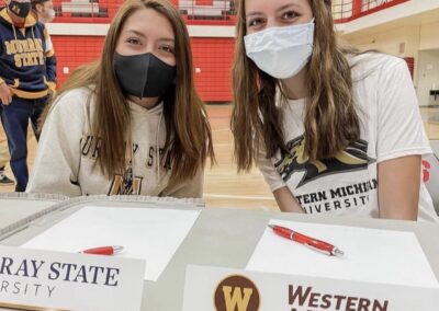 Two girls sit at a table with masks on their faces, celebrating college signing day.