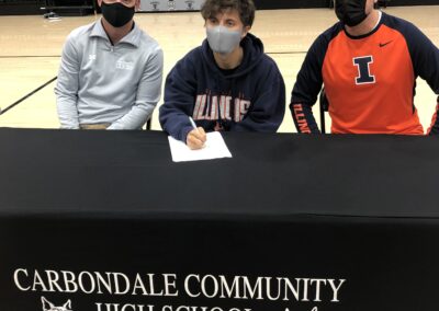 Three men sitting at a table during college signing day, signing a letter of intent.