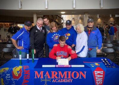 A group of people participating in a college signing day, standing around a table and signing a letter of intent.